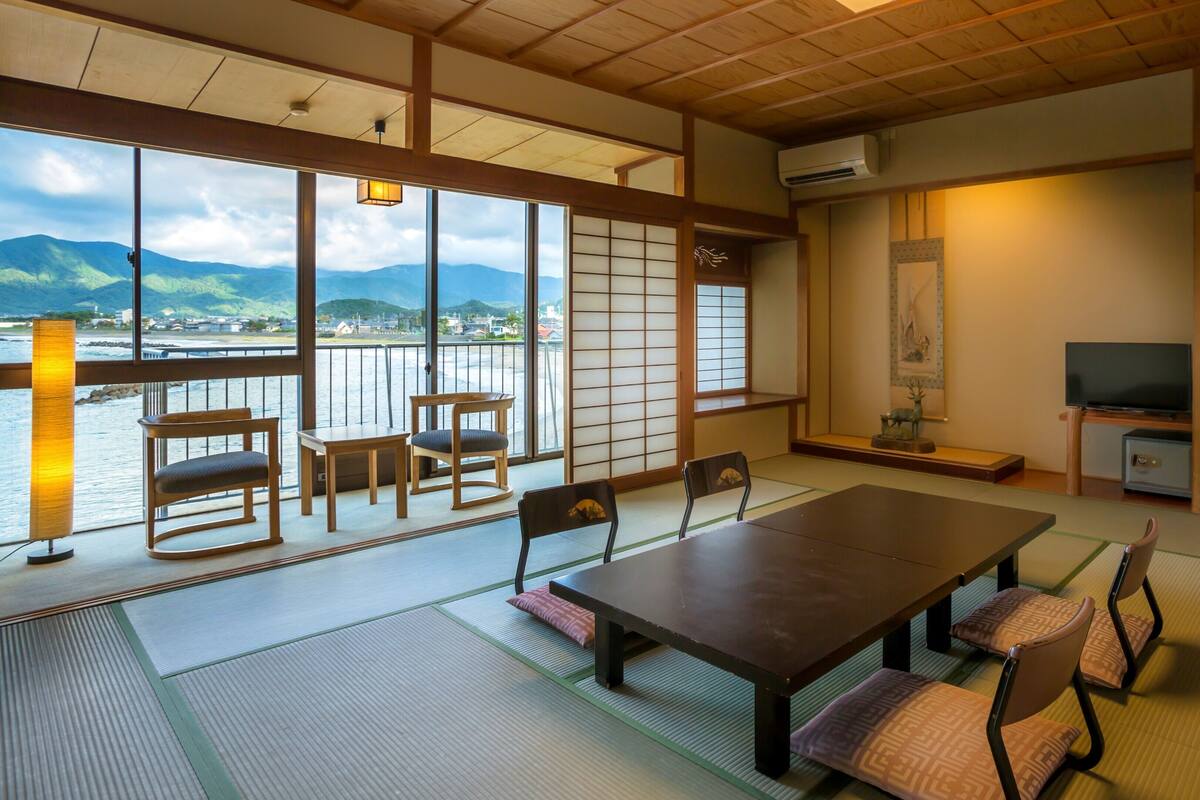 A traditional Japanese room with tatami flooring, a low table, chairs, a TV, and a view of mountains through large windows.