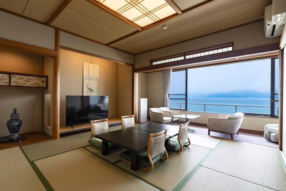 A modern hotel room with tatami flooring, a dining table, chairs, a TV, and a view of the sea.