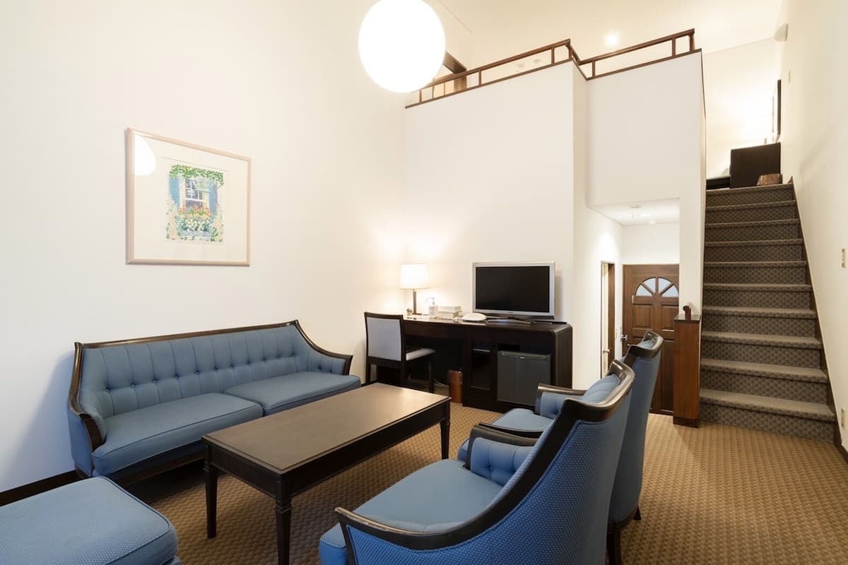 Habitación de hotel con sofá, sillones, una mesa de centro, televisión, escritorio y escalera.