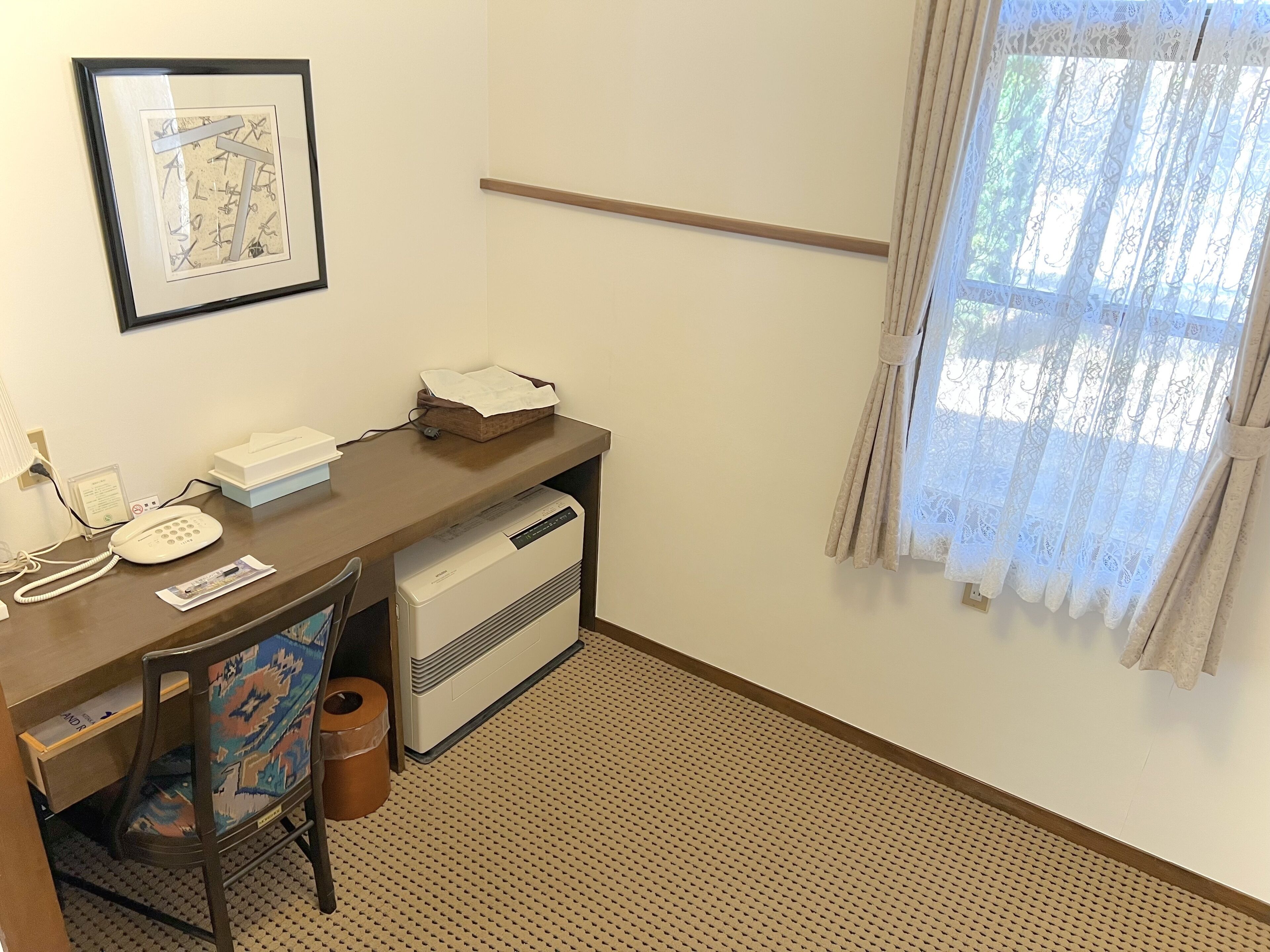 Une chambre d’hôtel avec un bureau en bois, un téléphone, une imprimante, une chaise, une poubelle, un tableau encadré au mur et une fenêtre avec des rideaux.