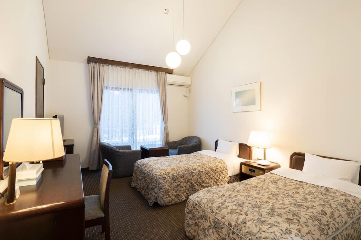 Habitación de hotel con dos camas, un escritorio, una silla, una mesita y un ventanal con cortinas.