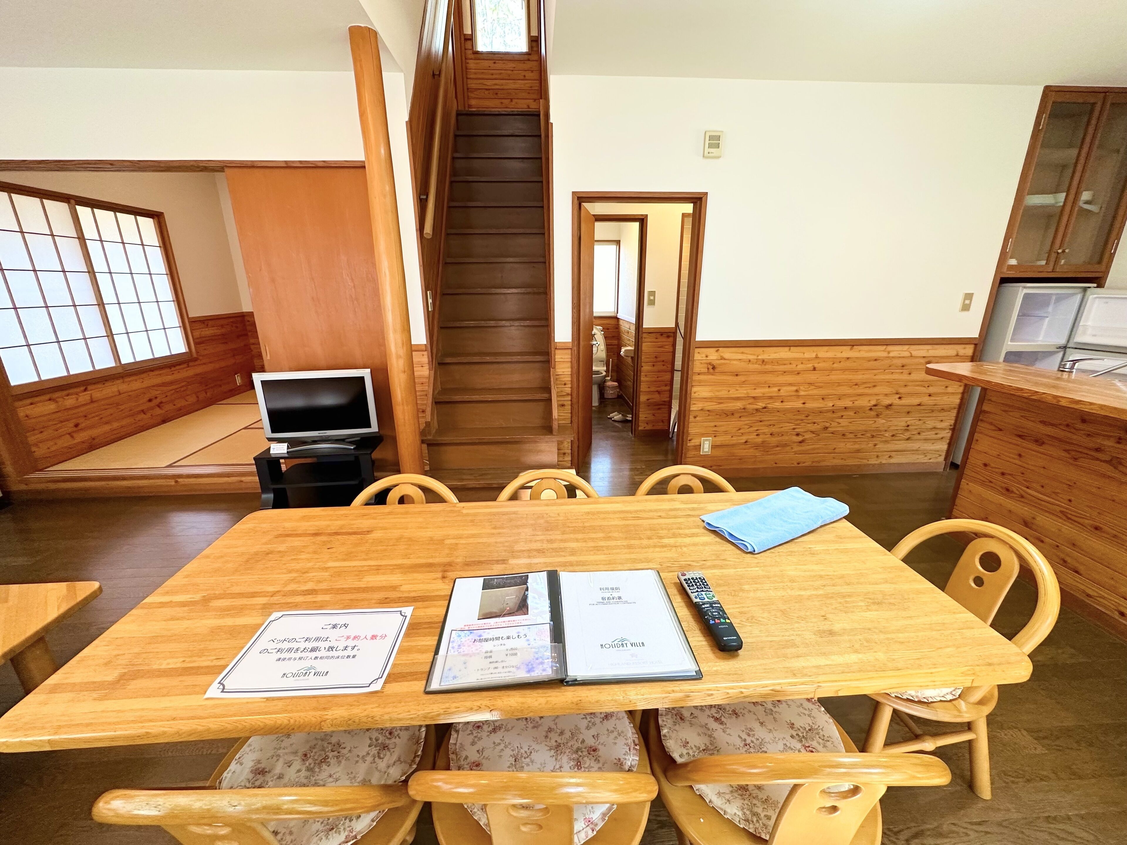 A traditional Japanese-style room with tatami flooring, a wooden dining table with chairs, a television, and a staircase leading to another level.
