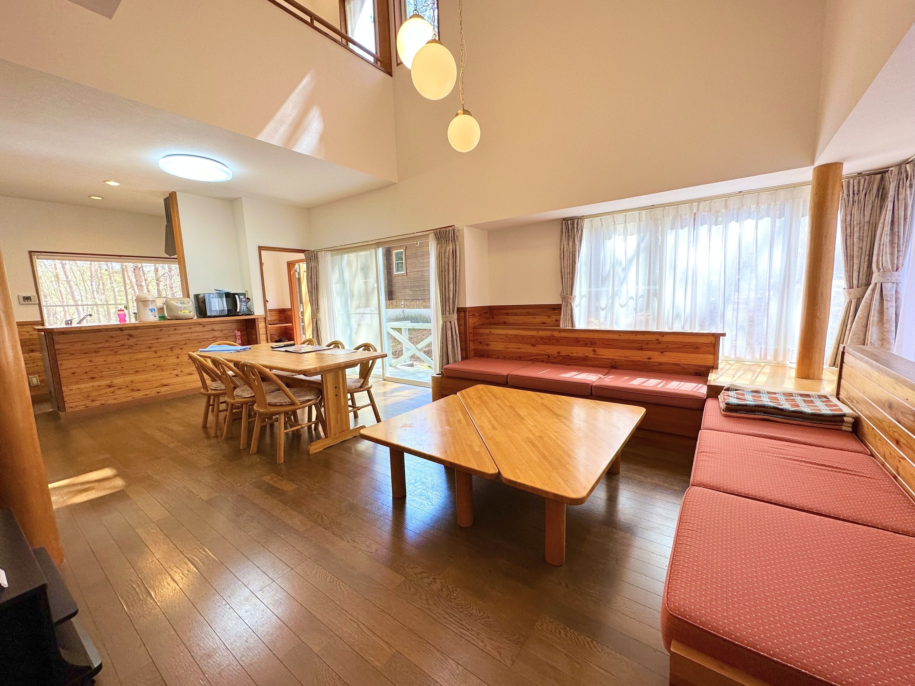 A spacious living area with a wooden dining table, a sofa, and a bench. There is a kitchen area with a counter and appliances. The room has large windows with curtains and a view of the outdoors.