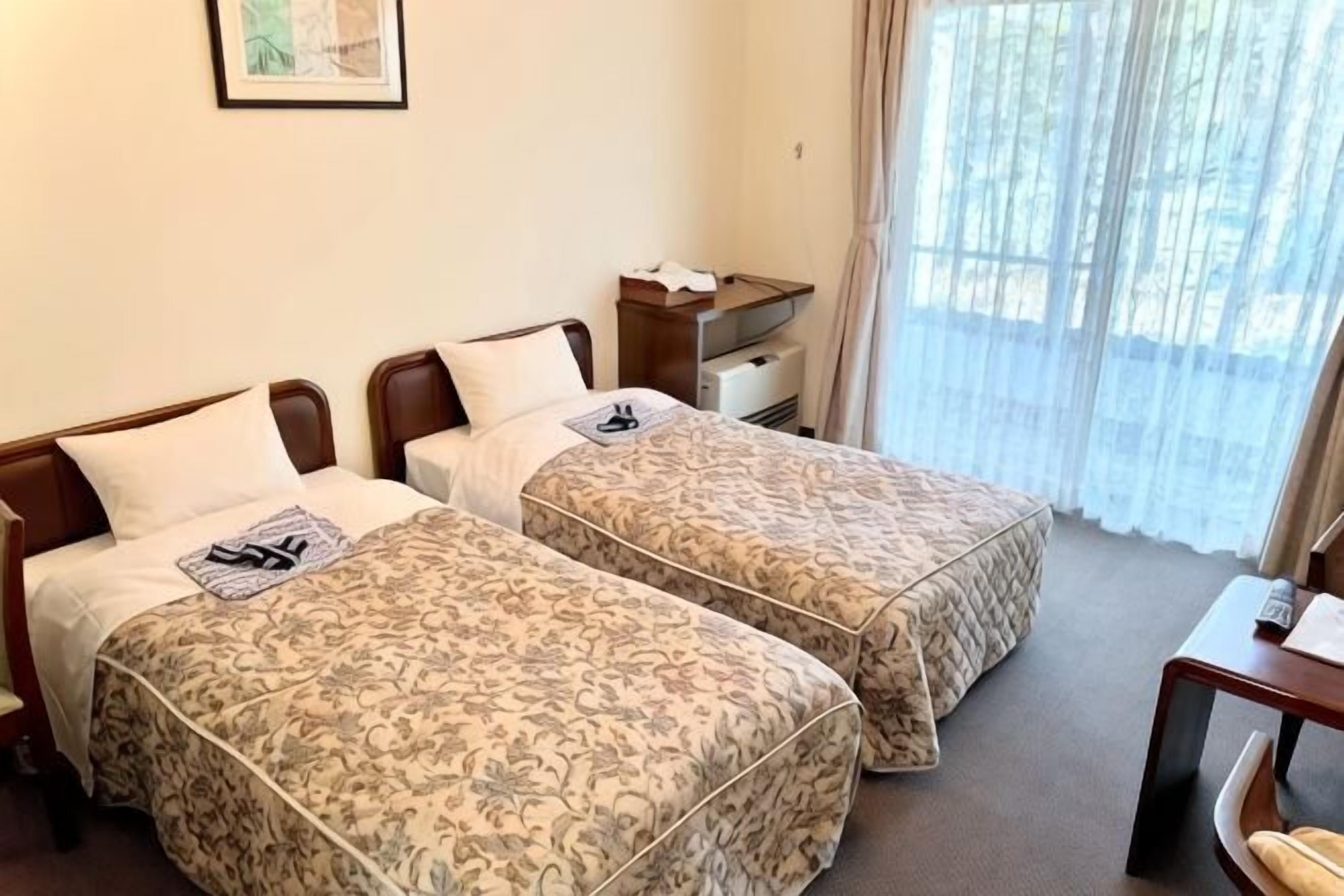 Hotellihuone, jossa on kaksi sänkyä, yöpöytä, työpöytä ja ikkuna, jossa on verhot.