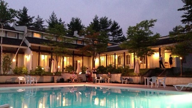 An outdoor swimming pool with lounge chairs and umbrellas, surrounded by buildings with warm lighting. - Holiday Villa Hotel & Resort Karuizawa (Tsumagoi)
