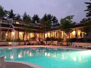 An outdoor swimming pool with lounge chairs and umbrellas, surrounded by buildings with warm lighting. - Holiday Villa Hotel & Resort Karuizawa (Tsumagoi)