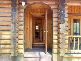 Una cabaña de madera con puerta central, un porche con mesa y sillas, y una ventana con vistas al bosque circundante.