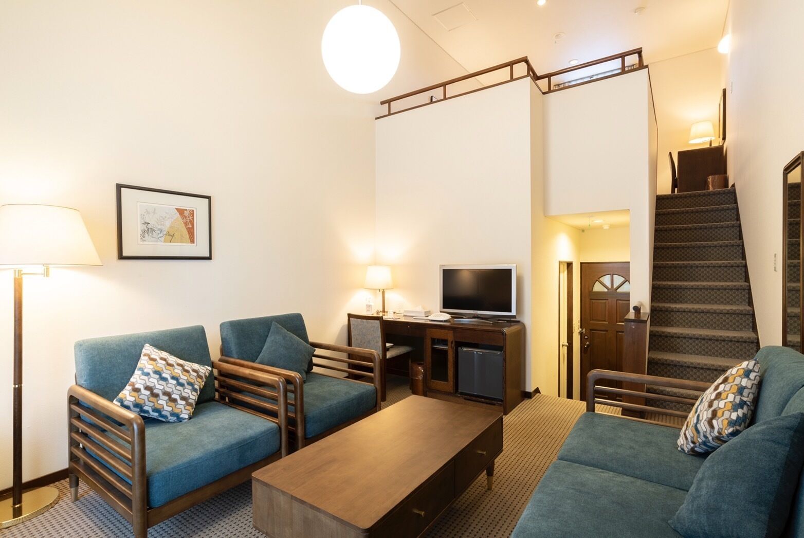 A hotel room with two blue sofas, a wooden coffee table, a flat-screen TV, a desk, and a staircase.