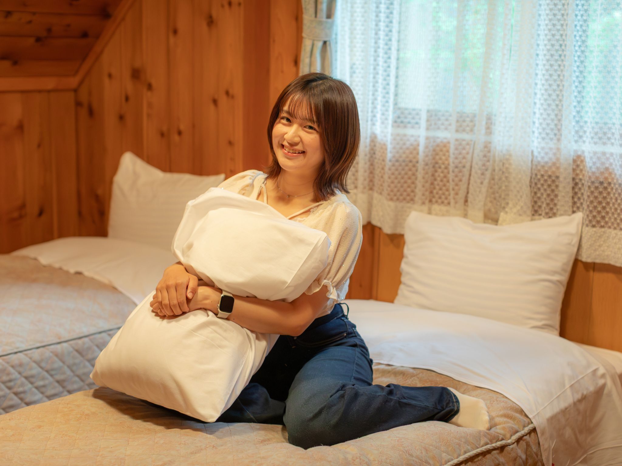 A person sitting on a bed in a room with wooden walls and a window with sheer curtains.