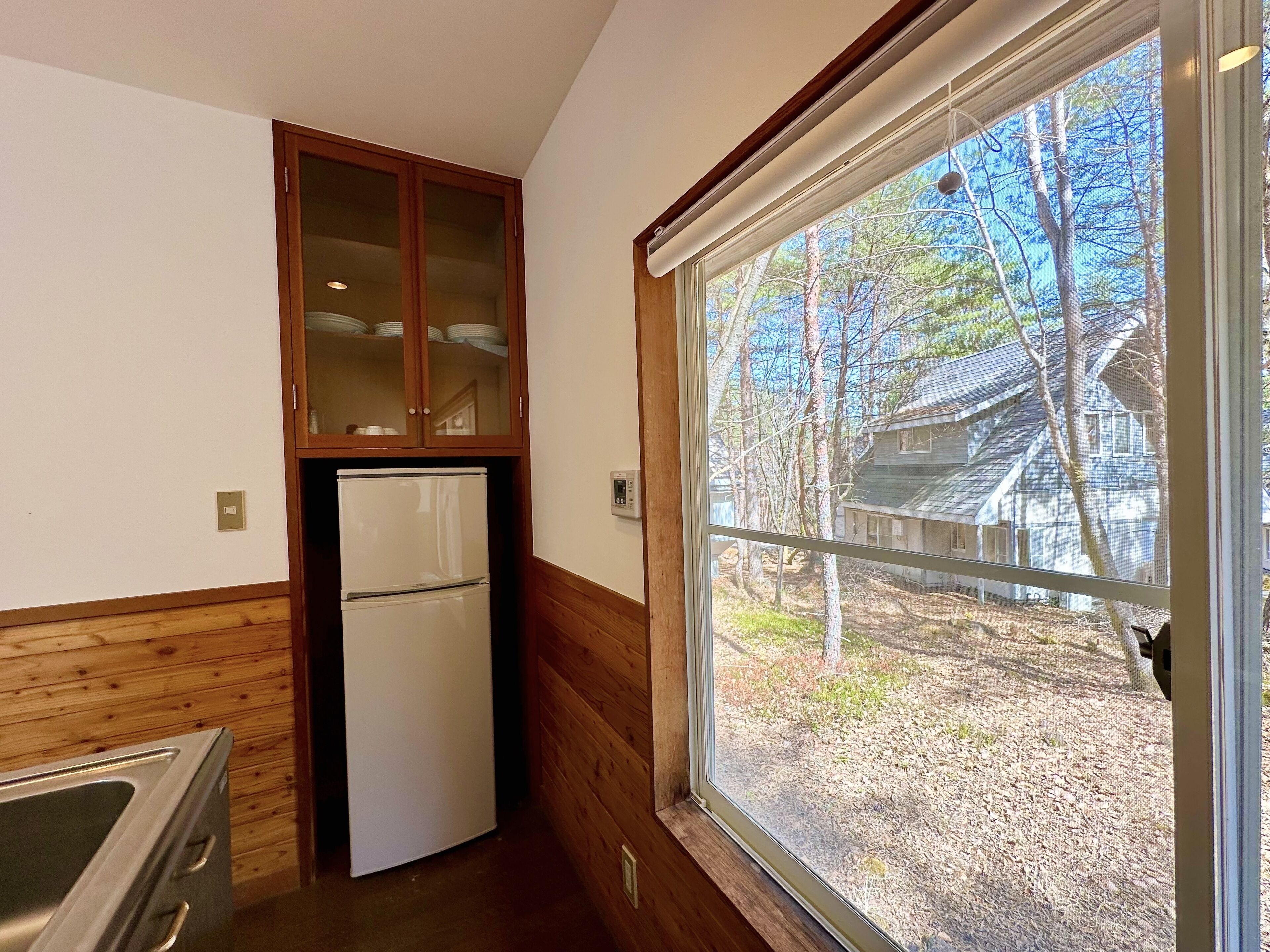 A compact kitchen with wooden paneling, a built-in refrigerator, and a window offering a view of a wooded area.
