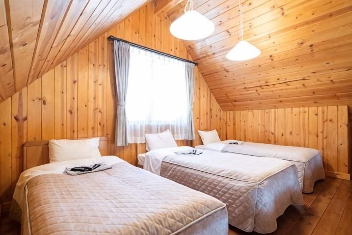 A room with wooden walls and sloped ceilings, two beds with white linens, a window with curtains, and hanging light fixtures.