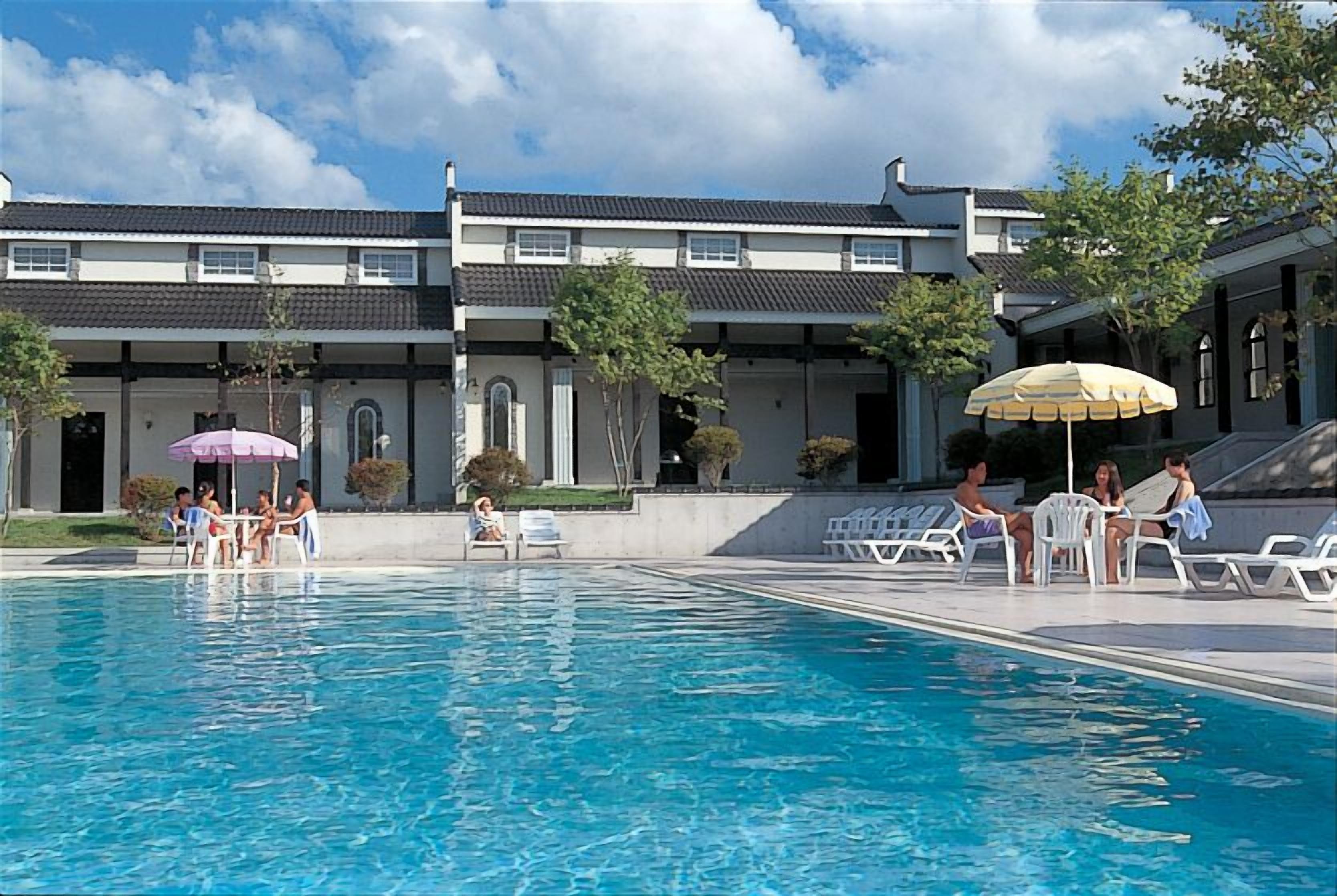 Area piscina con sedie a sdraio e ombrelloni, circondata da un edificio con tetto in tegole e diverse finestre.