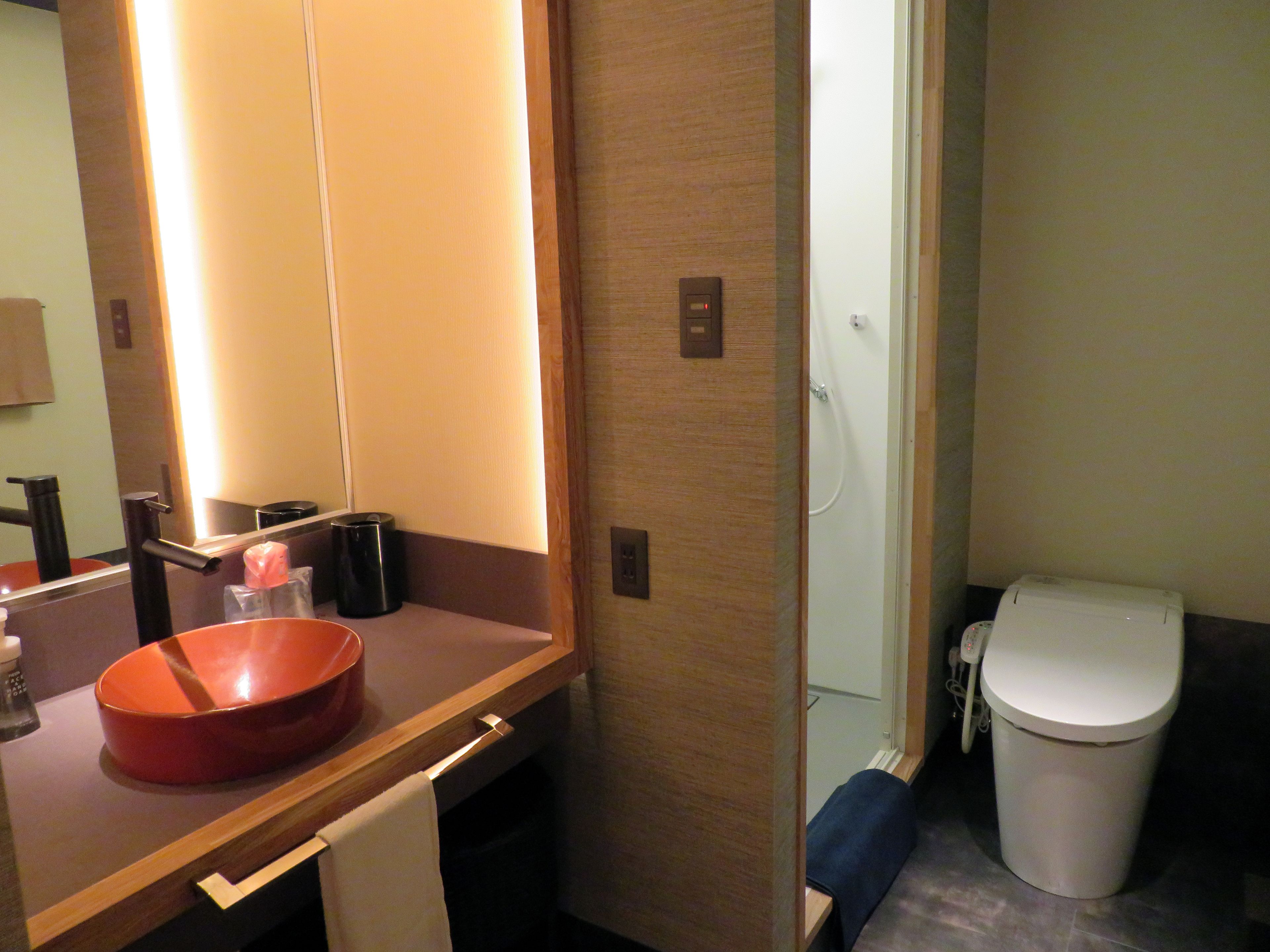 Un baño con tocador de madera, lavamanos, espejo, toalla, cubo de basura y retrete.