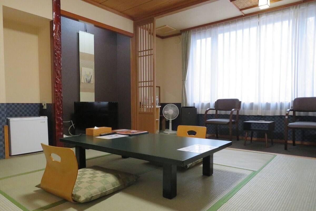 A traditional Japanese room with tatami flooring, a low table, chairs, a fan, and a television.