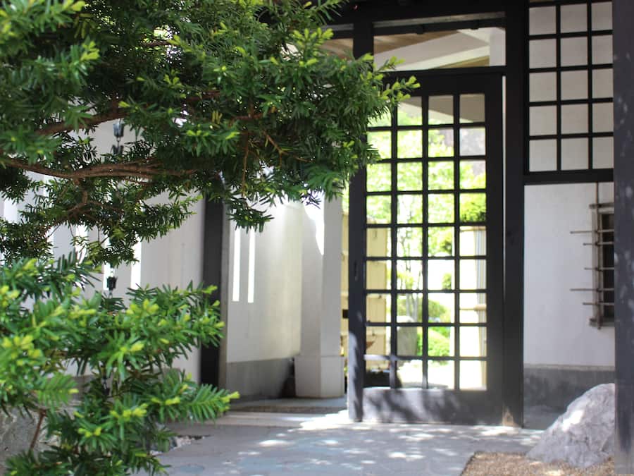 A traditional Japanese gate with a wooden frame and a grid pattern, leading to a building with white walls and a tiled floor.