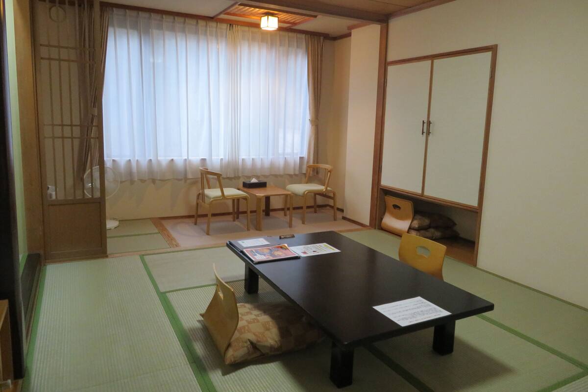 A traditional Japanese room with tatami flooring, a low table, two chairs, a cabinet, and a window with curtains.