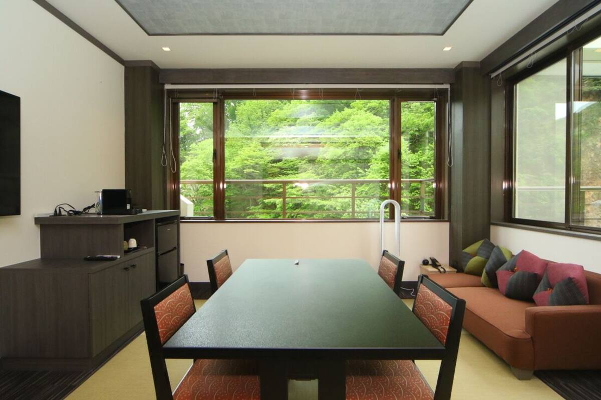 A modern room with a dining table, chairs, a sofa, a TV, and a window with a view of greenery.