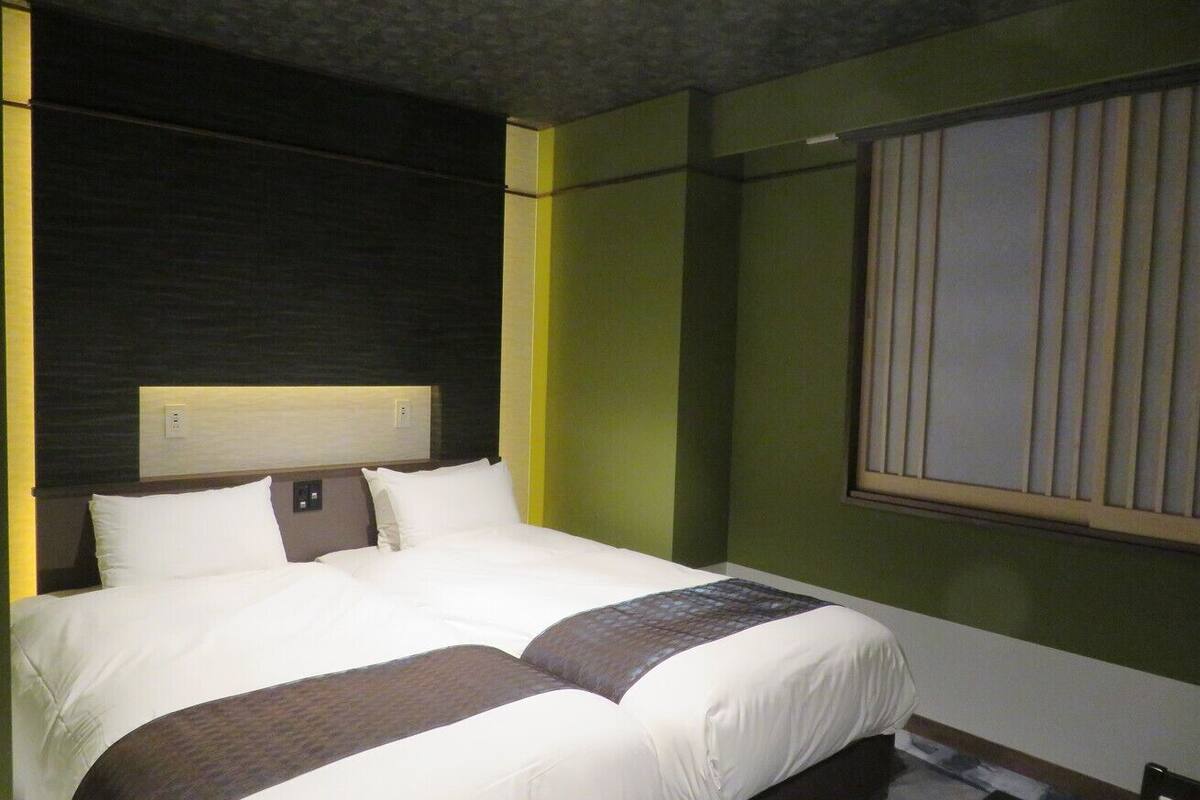 A hotel room with a large bed, a headboard with a light fixture, a window with blinds, and a green accent wall.