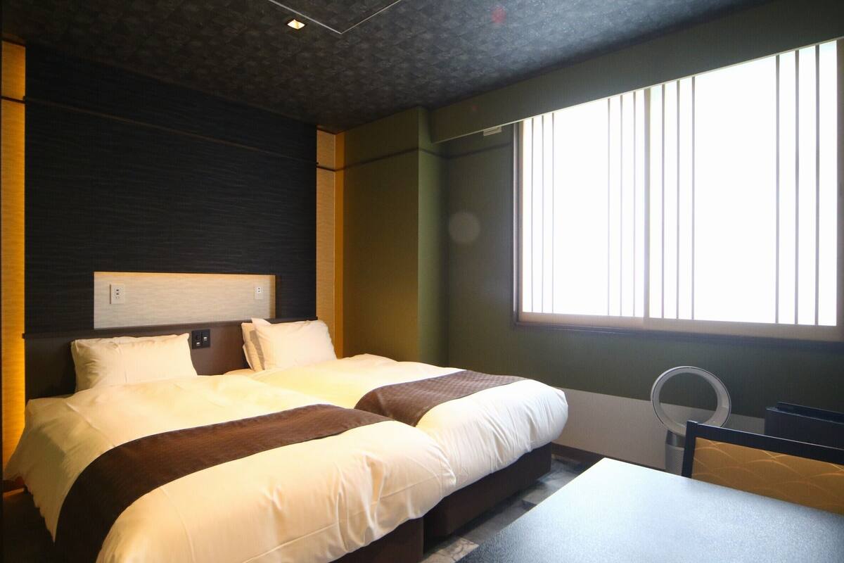 A hotel room with a large bed, a bedside table, and a window with blinds.
