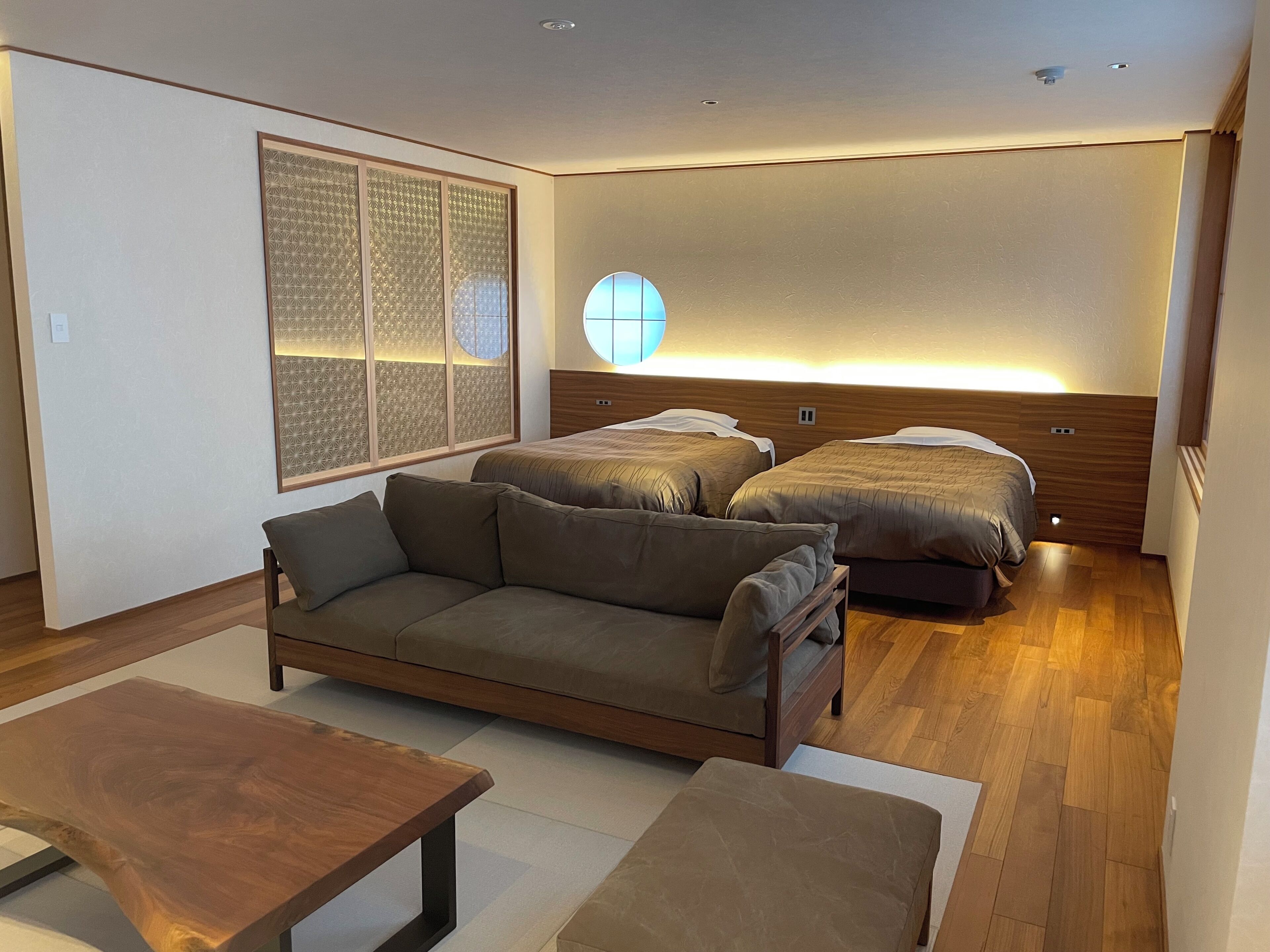 A modern bedroom with a sofa, two beds, a wooden headboard, and a large window with a geometric pattern.