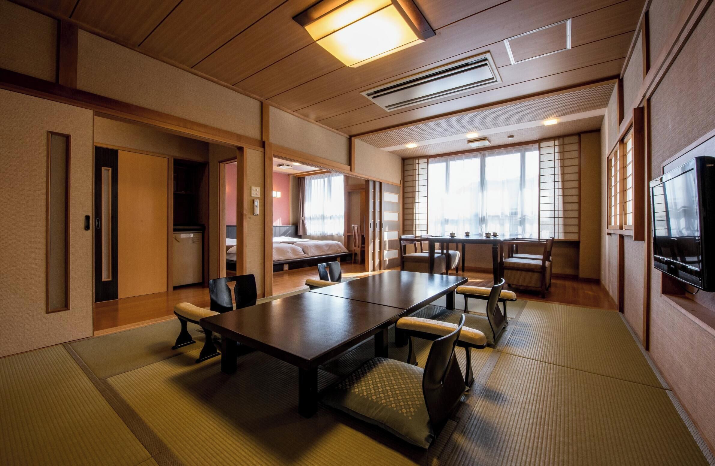 A hotel room with a large wooden dining table, chairs, a bed, a TV, and a view of the city.