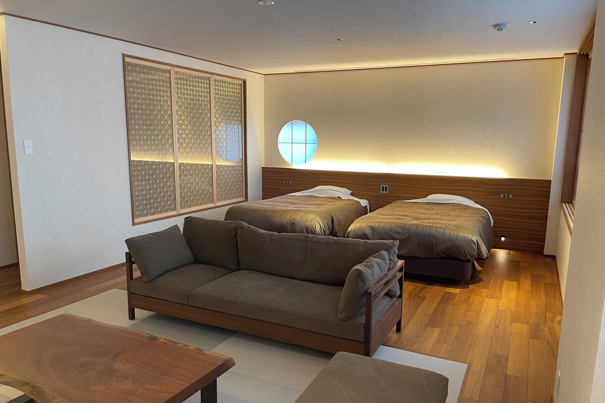 A modern bedroom with a sofa, two beds, a wooden headboard, and a large window with a geometric pattern.