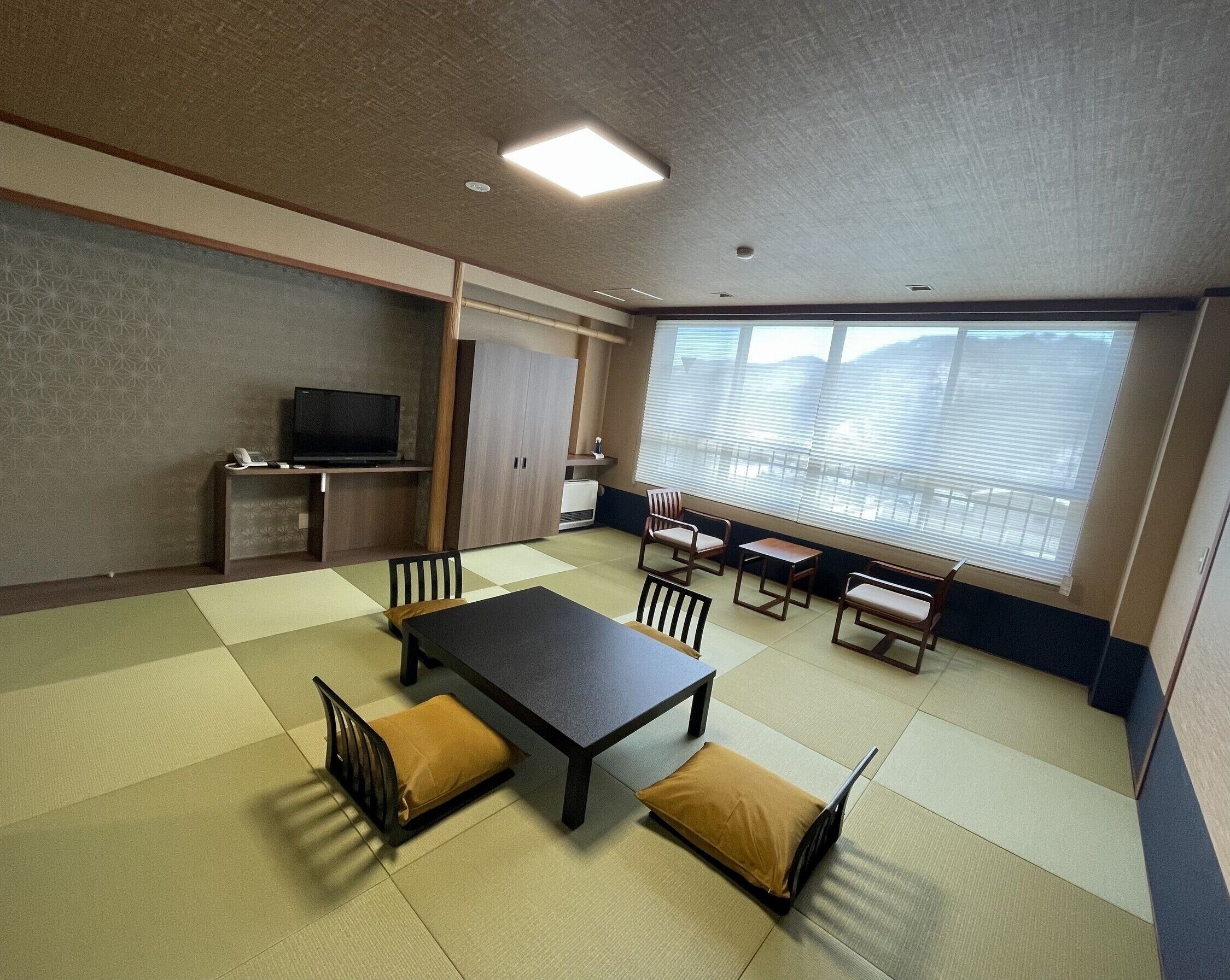 A room with tatami flooring, a tatami mat table, chairs, a television, and a large window with blinds.