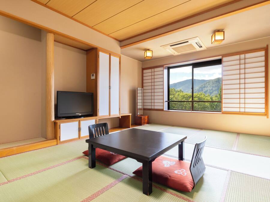 A traditional Japanese room with tatami flooring, a low table, chairs, a television, sliding doors, and a view of mountains.