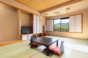 Una habitación japonesa tradicional con suelo de tatami, una mesa baja, sillas, televisión, puertas correderas y vistas a montañas.