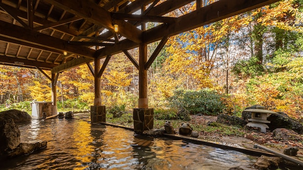 Un onsen japonés tradicional con vigas de madera y faroles de piedra, rodeado de follaje otoñal.