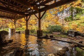 Un onsen japonais traditionnel, avec des poutres en bois et des lanternes en pierre, entouré d’un feuillage automnal.