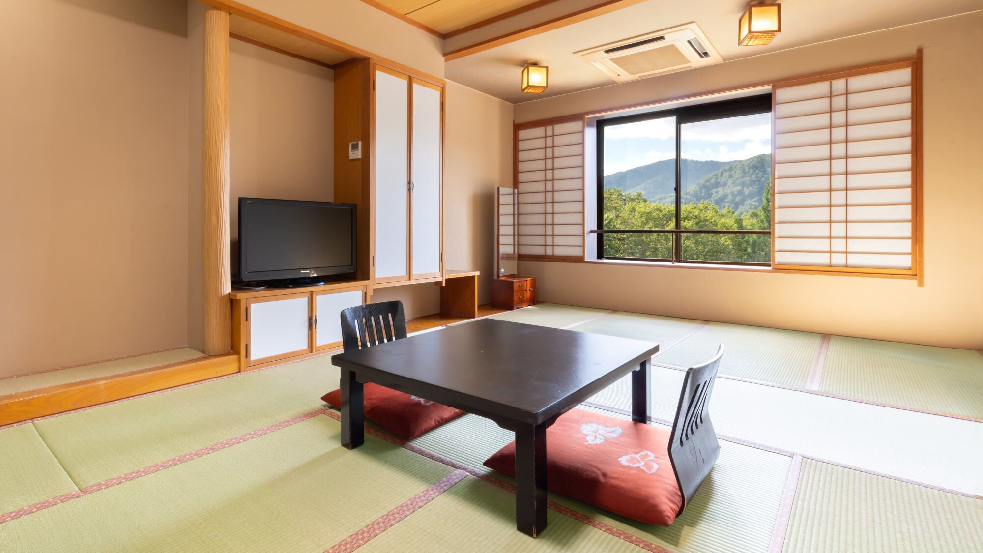 Une pièce de style japonais traditionnel, avec un sol recouvert de tatamis, une table basse, un téléviseur, des portes coulissantes et une vue sur une montagne boisée.
