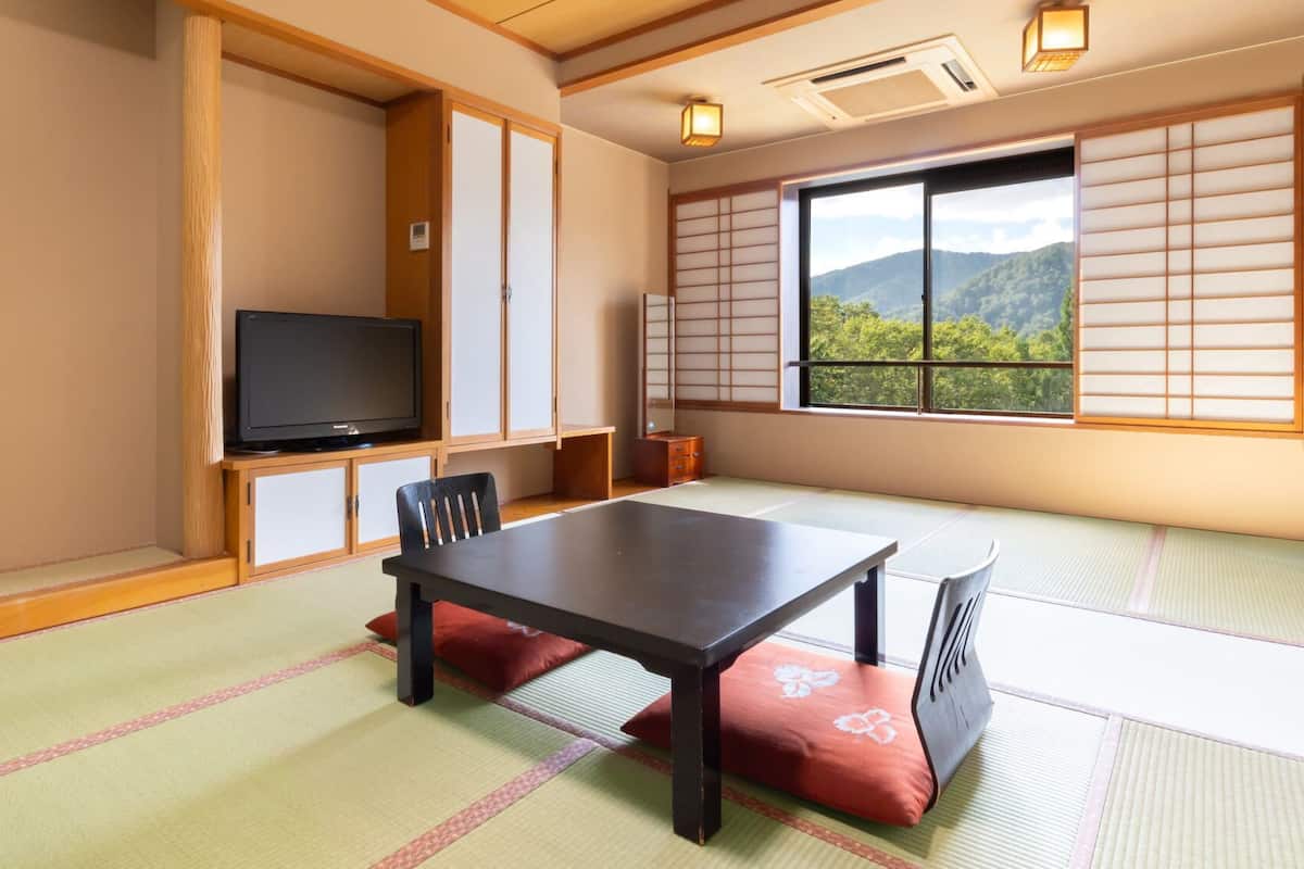 Une pièce de style japonais traditionnel, avec un sol recouvert de tatamis, une table basse, un téléviseur, des portes coulissantes et une vue sur une montagne boisée.