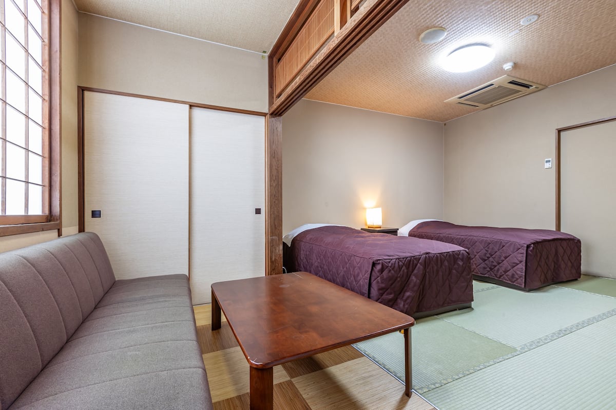 Une pièce de style japonais traditionnel, avec un sol recouvert de tatamis, une basse table en bois, un banc rembourré et deux lits avec une literie violette.