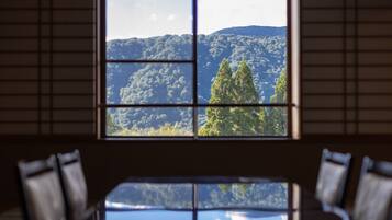 A room with a large window divided into four panes, offering a view of a forested mountain. There are two chairs in the foreground.