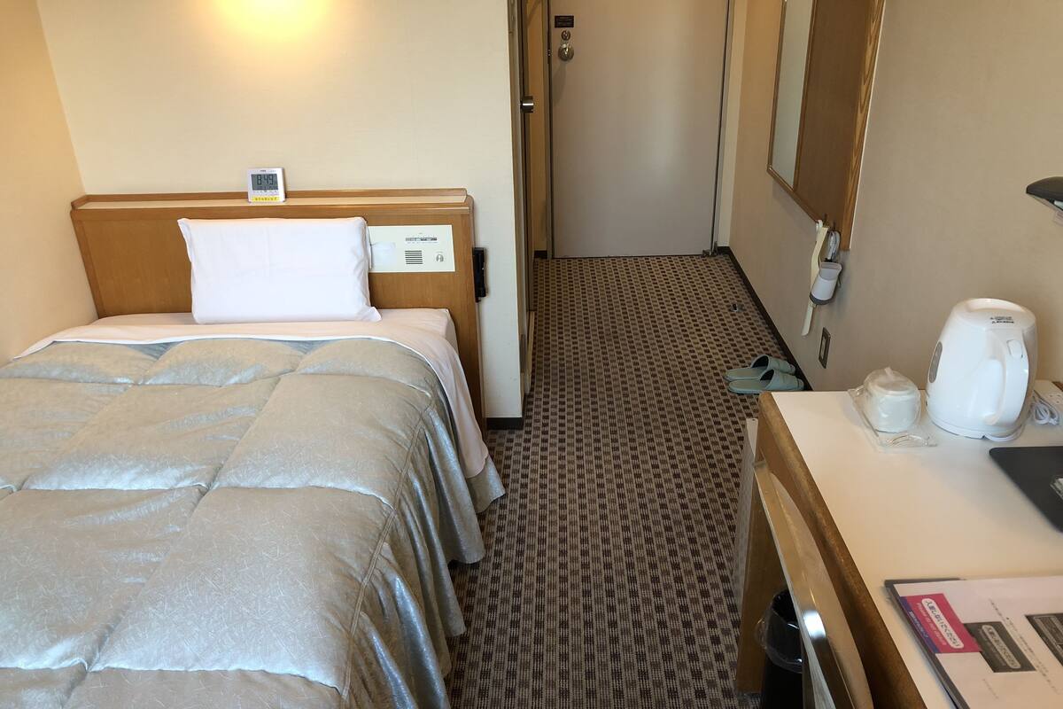 A hotel room with a bed, bedside table, and a small desk with a kettle and a cup.