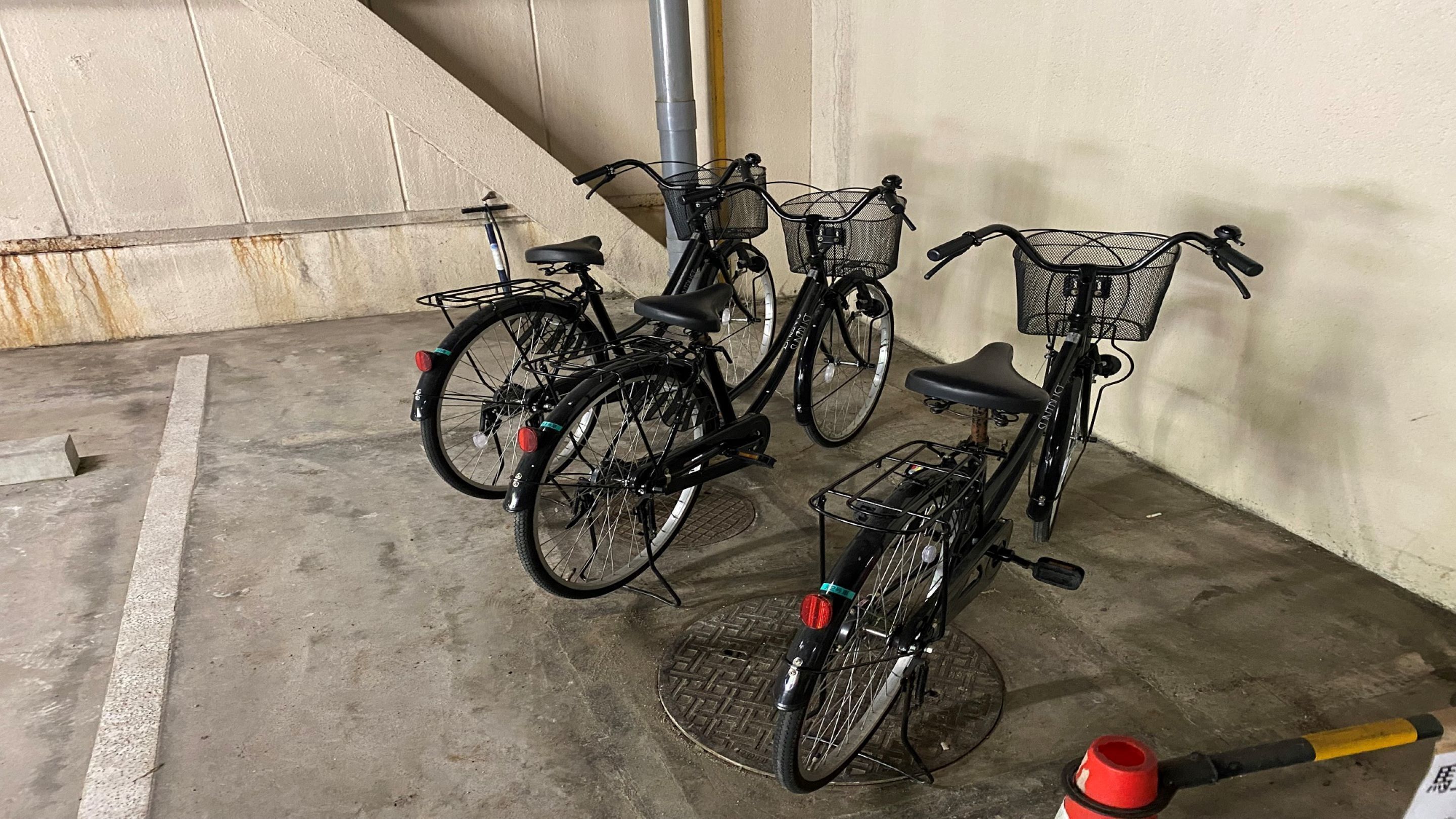 A parking area with bicycles parked against a wall, a metal grate on the floor, and a red and yellow traffic cone.