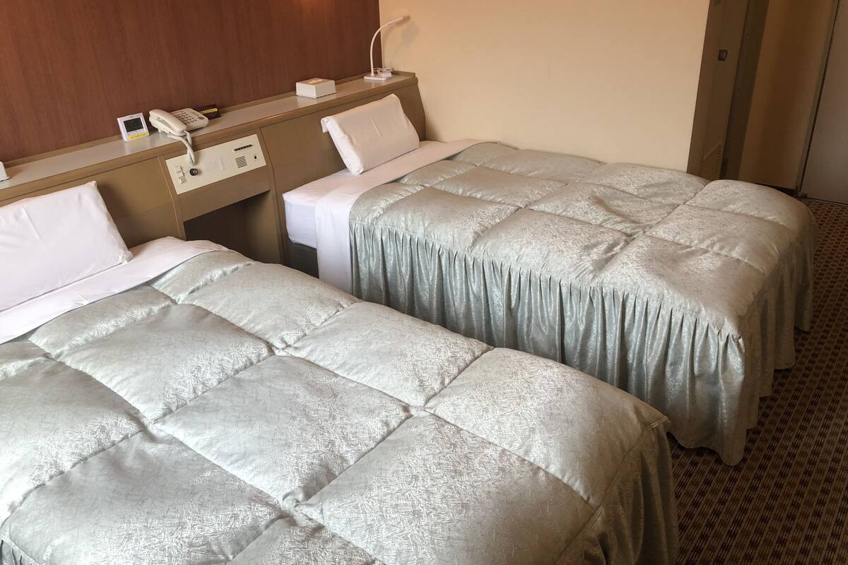 A hotel room with two beds, a bedside table with a phone and a digital clock, and a wall-mounted lamp.