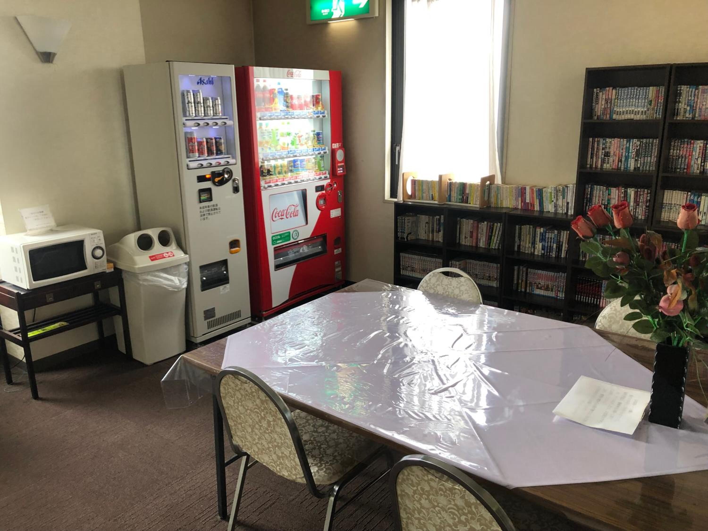 A room with a table covered in plastic, vending machines, a microwave, and a bookshelf filled with books.