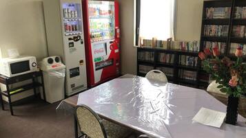 A room with a table covered in plastic, vending machines, a microwave, and a bookshelf filled with books.