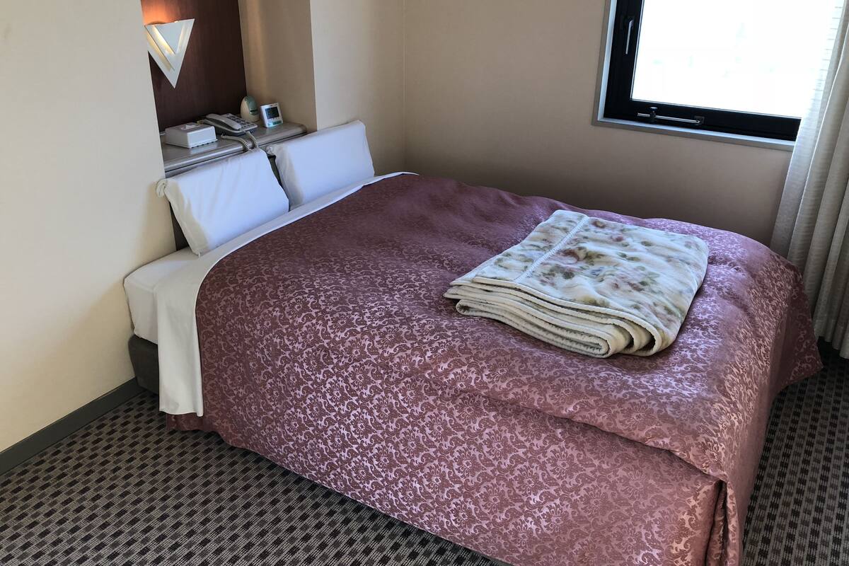 A hotel room with a bed, a bedside table with a phone and a small device, a wall-mounted lamp, and a window with curtains.