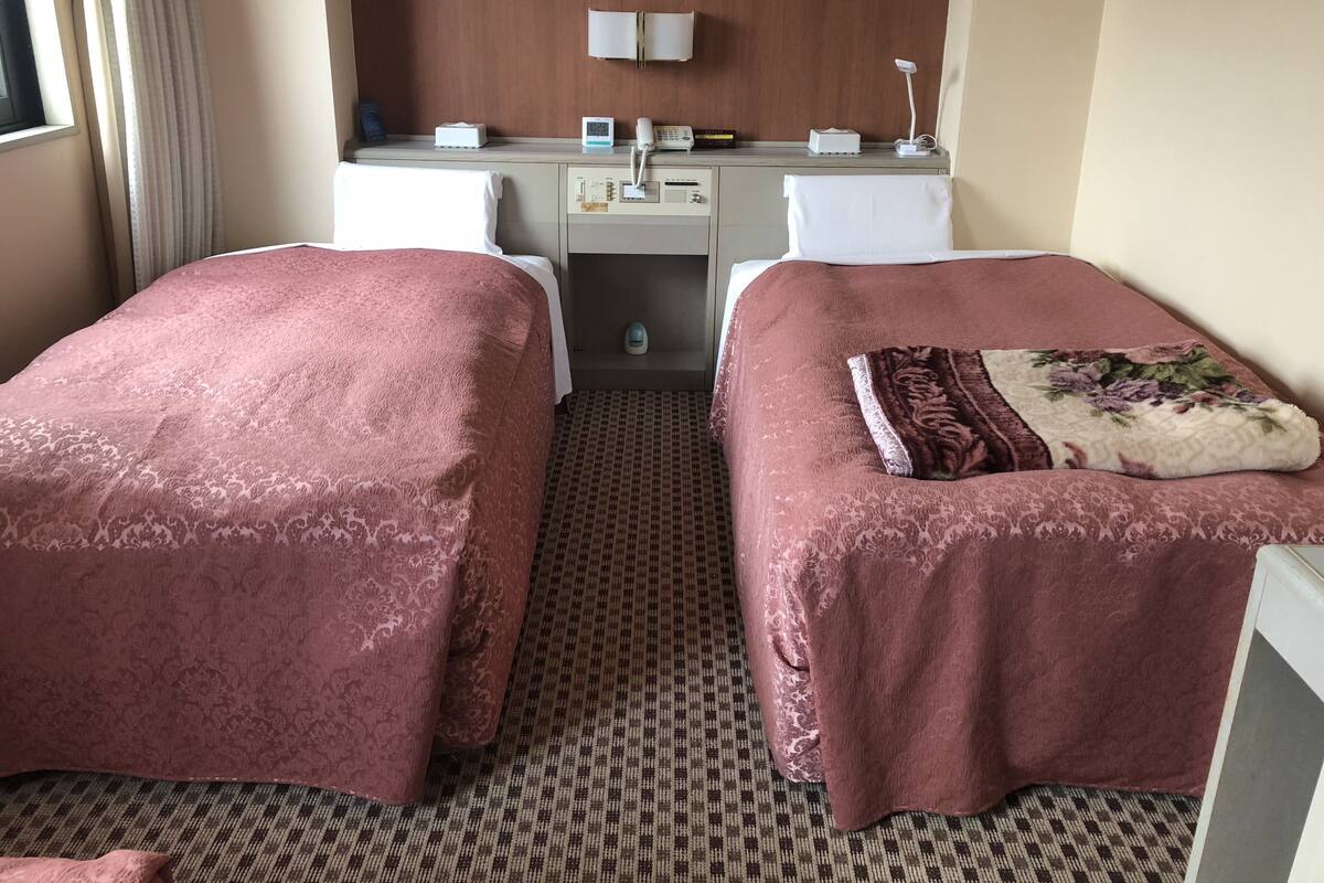 A hotel room with two beds, a desk, a telephone, and a small window with curtains.