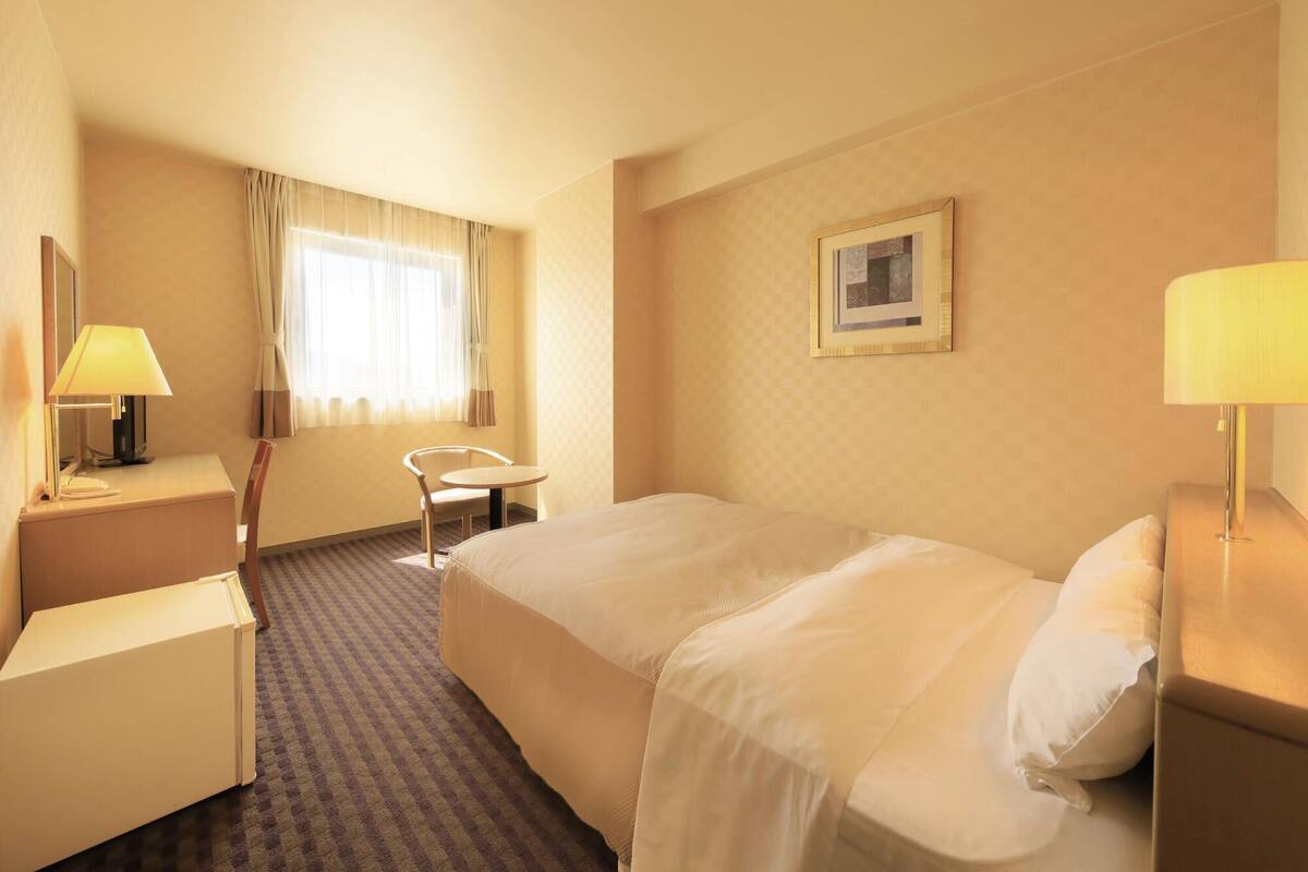 Een hotelkamer met een bed, bureau, stoel, lamp en een raam met gordijnen.
