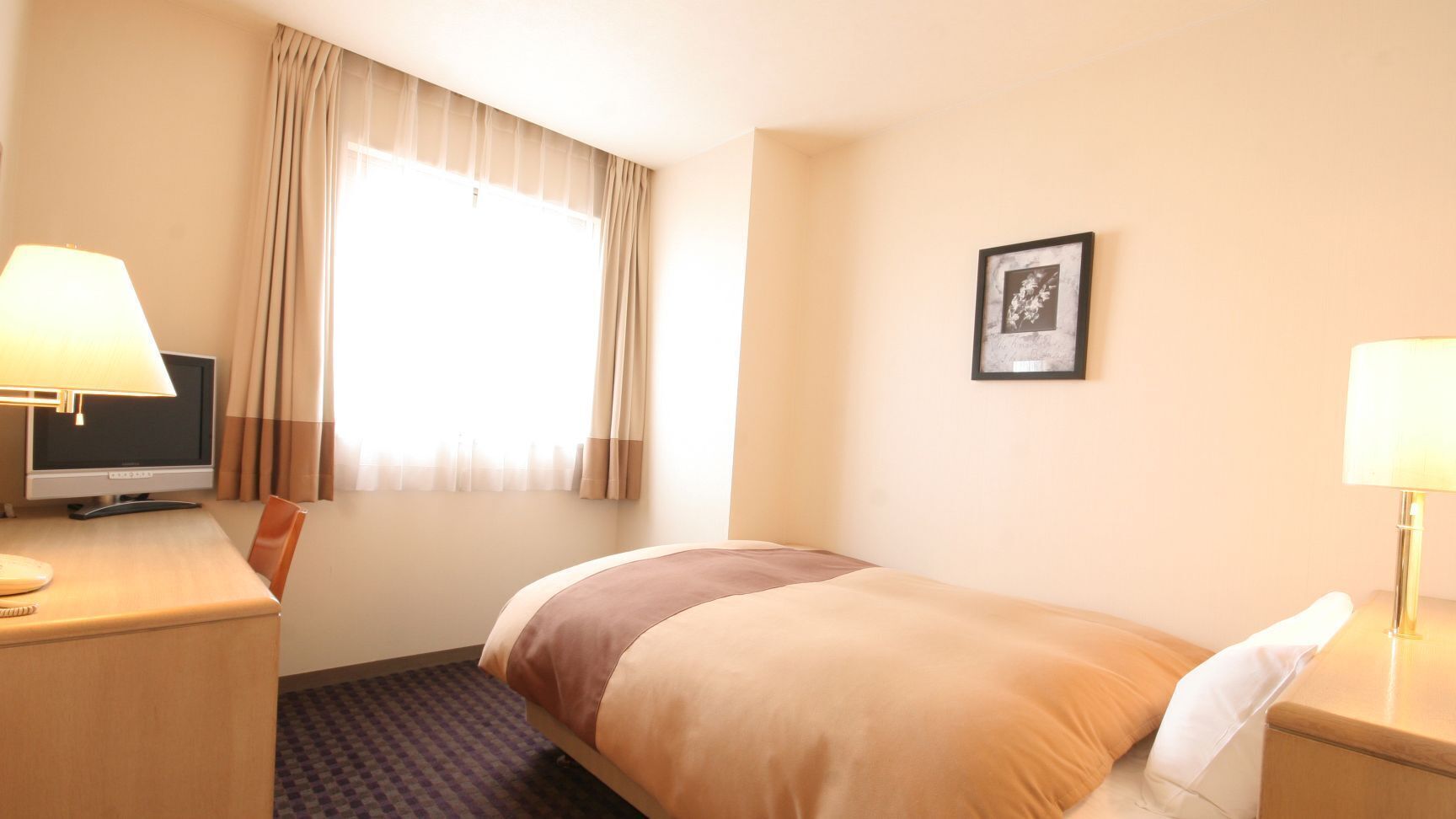 Een hotelkamer met een bed, een bureau met een computer, een stoel, een raam met gordijnen en een lijst met een afbeelding aan de muur.