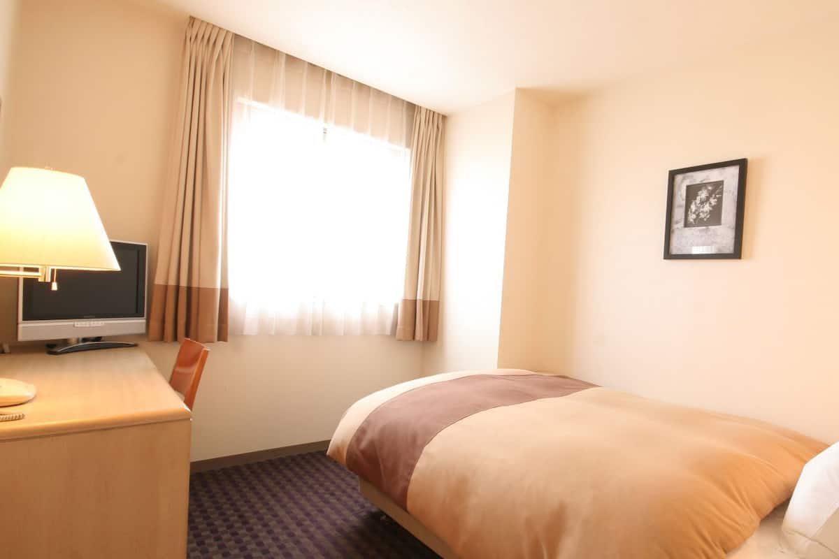 A hotel room with a bed, a desk with a computer, a chair, a window with curtains, and a framed picture on the wall.