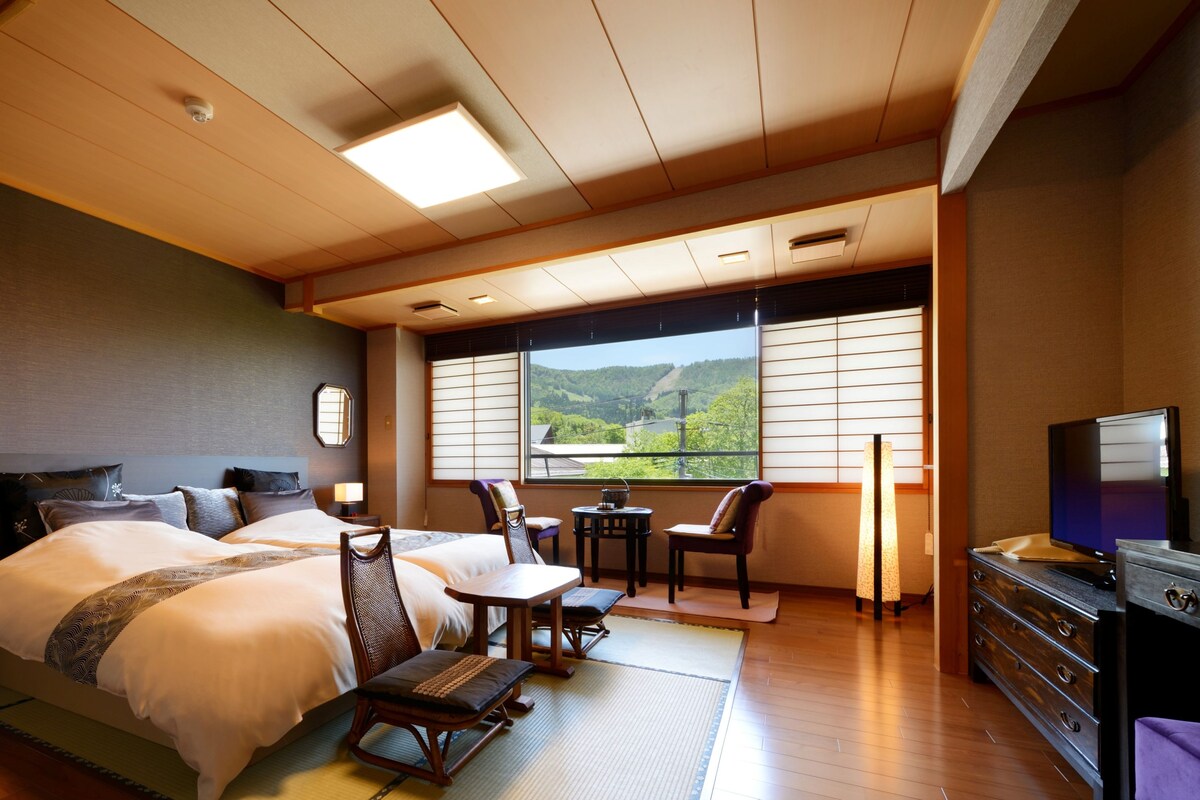 A hotel room with a bed, two chairs, a desk, a TV, and a view of a mountain.