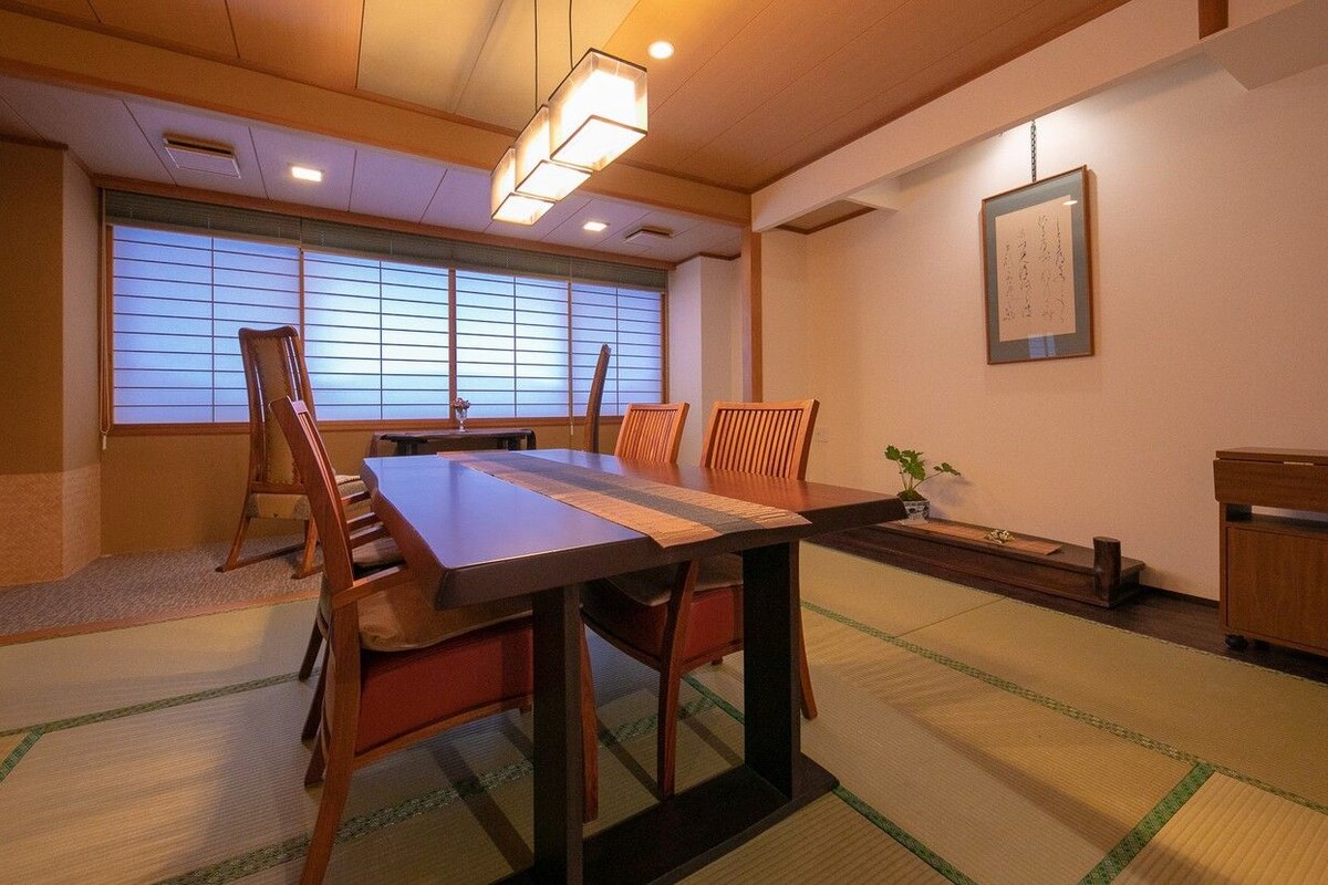 A traditional Japanese room with tatami flooring, a low table, wooden chairs, a small cabinet, and a window with shoji screens.