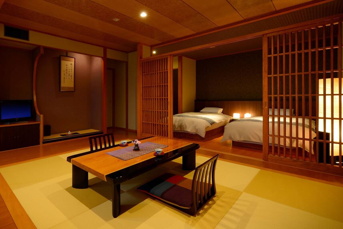 Ein traditionell japanischer Raum mit Tatami-Boden, einem niedrigen Tisch, einem Fernseher und einem Bett mit einem Holzbalkon.