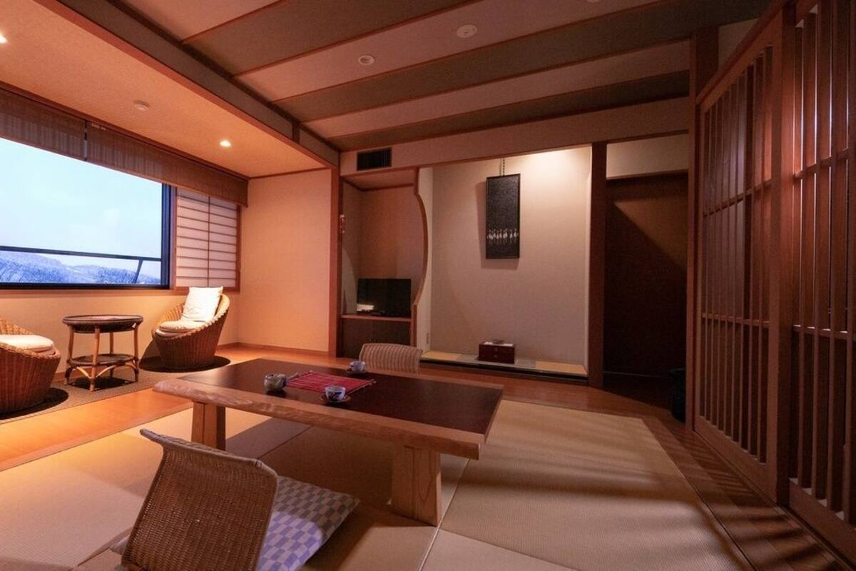 Ein traditioneller japanischer Raum mit Tatami-Böden, einem niedrigen Tisch, zwei gepolsterten Stühlen, einem Fernseher und einer Schiebetür.