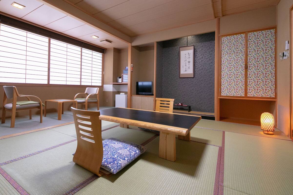 Ein traditioneller japanischer Raum mit Tatami-Boden, einem niedrigen Tisch, Holzstühlen, einem Fernseher und einem Shoji-Bildschirm.